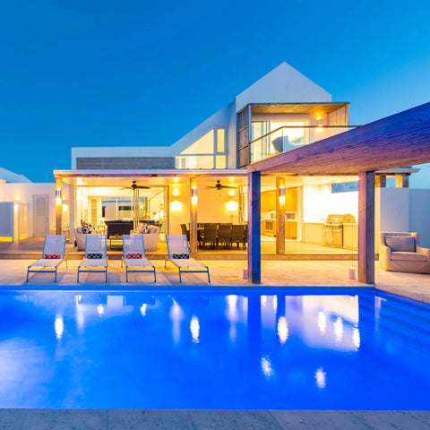 Contemporary villa at dusk with illuminated pool, lounge chairs, and outdoor dining area. Modern windows and architecture feature the Flores Sconce by A19 Artisan Lighting—USA-made wall lighting, handcrafted in California.