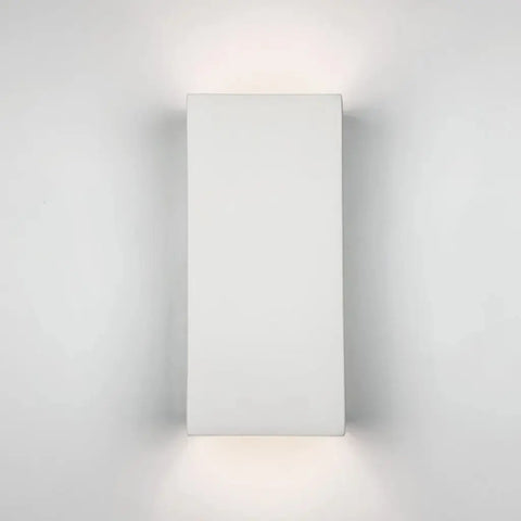 The Modern USA-Made Flores Sconce by A19 Artisan Lighting, handcrafted in California, features a minimalist rectangular design and custom finish, mounting on a white wall to cast soft up-and-down light. LED smart bulb compatible for efficiency.