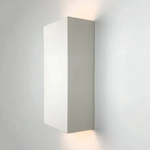 The Modern USA-Made Flores Sconce by A19 Artisan Lighting, handcrafted in California, is a rectangular white fixture that emits gentle upward and downward light and is compatible with LED smart bulbs for customizable illumination.