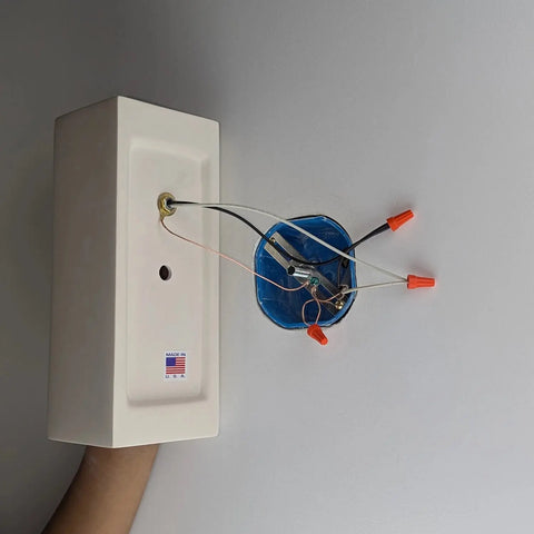 A hand holds the white Modern Flores Sconce by A19 Artisan Lighting near a circular electrical box with orange wire nuts against a light gray wall. Handcrafted in California, this USA-made wall light is ideal with LED smart bulbs and custom finishes available.