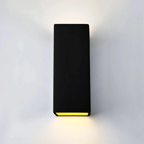 The Modern USA-Made Wall Lighting – Flores Sconce by A19 Artisan Lighting, handcrafted in California, features a sleek black rectangular design that emits soft yellow light both upward and downward, creating a gentle glow on a white wall.