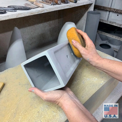 In a workshop, someone uses a yellow sponge to smooth a gray ceramic piece for the Modern USA-Made Flores Sconce, handcrafted in California by A19 Artisan Lighting. Tools are on the workbench and a MADE IN U.S.A. label appears in the corner.