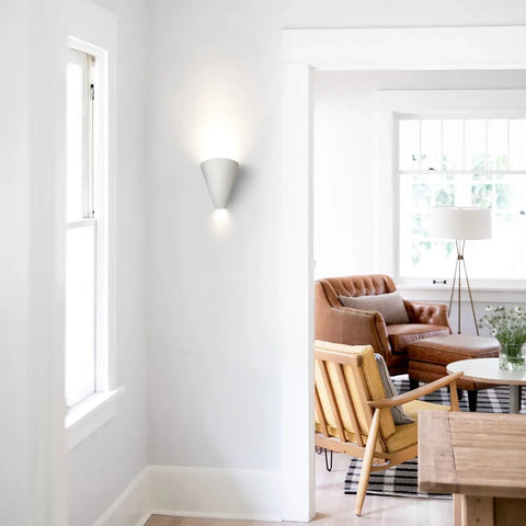 A modern, bright living space with a USA-made Gotlandia Sconce by A19 Artisan Lighting, energy-efficient LEDs, a wooden dining table, tan leather sofa, wood armchair, floor lamp, and large windows flooding the room with natural light.