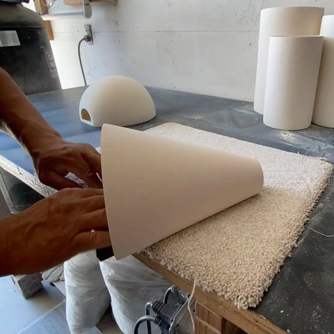 Hands smooth a cone-shaped ceramic piece on a workbench for A19 Artisan Lighting’s Modern USA-Made Gotlandia Sconce. Other unfinished ceramic shades and pottery tools highlight the handcrafted California process.
