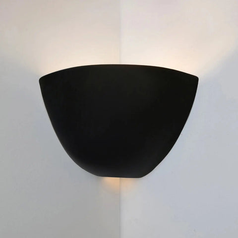 The Gran Aegina Corner Sconce by A19 Artisan Lighting, handcrafted in California, is a modern black fixture mounted on a white wall corner. It uses LED smart bulbs to cast soft up and down light, creating a gentle glow on both walls.