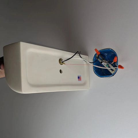 The Modern USA-Made Gran Balboa Downlight Sconce by A19 Artisan Lighting, handcrafted in California, is a beige wall fixture with exposed wires mounted sideways near an open blue box on a gray wall.