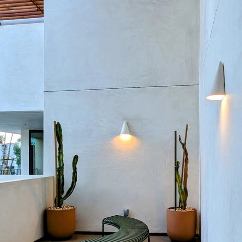 A modern minimalist balcony features two potted cacti, a curved green bench, and white textured walls illuminated by two Icelandia Sconces—handcrafted in California by A19 Artisan Lighting.