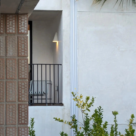 A small balcony with a metal chair and the Modern USA-Made Icelandia Sconce by A19 Artisan Lighting, handcrafted in California, is partly visible behind a brick wall. Green foliage grows in front as LED smart bulbs softly light the space.