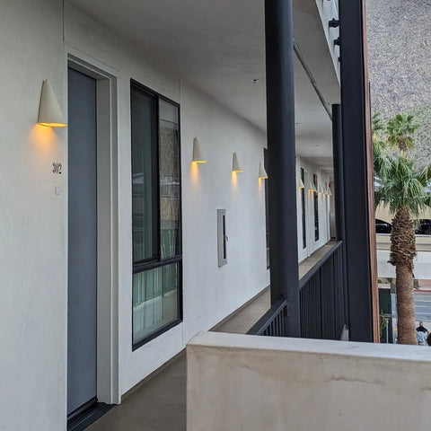 A modern hotel exterior features room doors and windows along a white corridor lit by Icelandia Sconce wall lights from A19 Artisan Lighting, handcrafted in California. Palm trees and parked cars appear in the background.