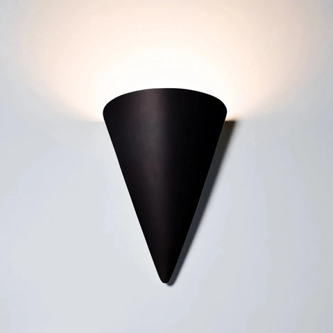 The Icelandia Sconce by A19 Artisan Lighting is a USA-made, wall-mounted black sconce handcrafted in California that casts warm upward light with optional LED smart bulbs, creating a soft glow on the white wall above.