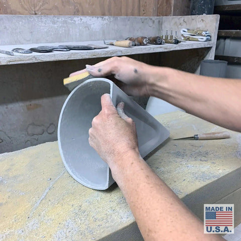 In a workshop, someone sands the edge of a ceramic bowl as the Modern USA-Made Wall Lighting – Icelandia Sconce by A19 Artisan Lighting is seen in the background, with a Made in U.S.A. label visible in the corner.