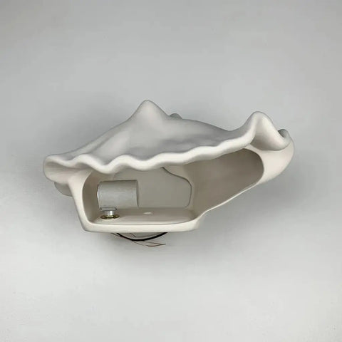 The Key West Sconce by A19 Artisan Lighting is a white, shell-shaped ceramic wall fixture, handcrafted in California and compatible with LED smart bulbs, shown mounted on a plain white wall with exposed wires and an empty socket.