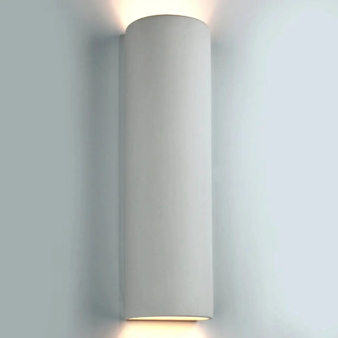 The Modern USA-Made Kos Sconce by A19 Artisan Lighting features a sleek cylindrical design, emitting soft light from both ends and casting a gentle glow on the wall. Handcrafted in California for contemporary spaces.