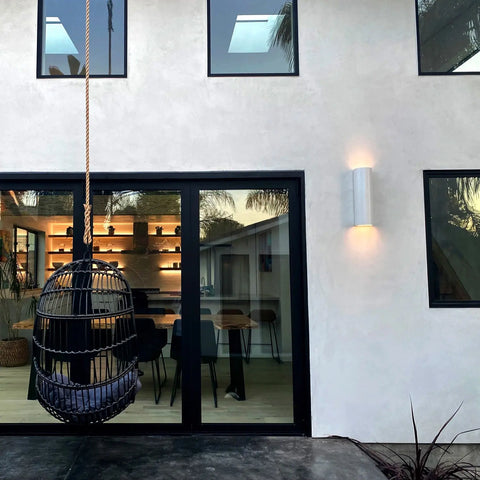 A modern house exterior with a large glass sliding door, black-framed windows, a hanging wicker chair with cushion, and warm light from A19 Artisan Lighting’s USA-made Kos Sconce; shelves and bar stools can be seen inside.