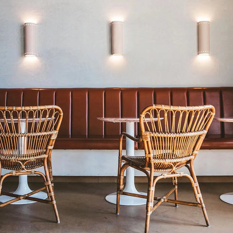 Two empty wicker chairs sit at round tables in front of a brown cushioned bench, illuminated by three Modern USA-Made Wall Lighting – Kythnos Sconce fixtures by A19 Artisan Lighting on the minimalistic café’s white wall.