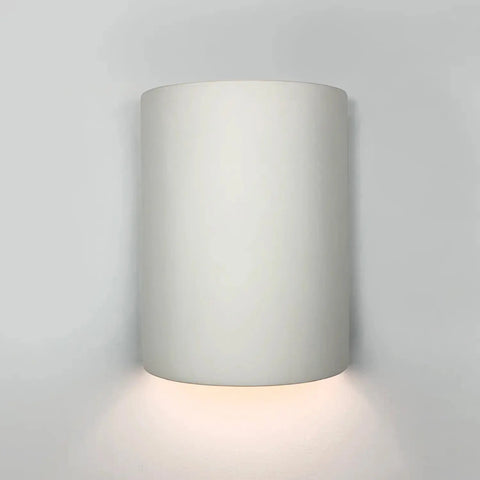The A19 Artisan Lighting Leros Downlight Sconce, handcrafted in California, features a modern cylindrical white design and emits a soft LED glow downward against a light grey wall.