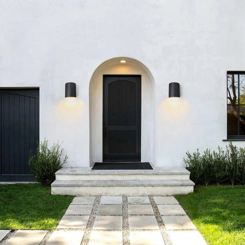A modern house entrance features a black door under an archway, flanked by two Leros Downlight Sconces from A19 Artisan Lighting, handcrafted in California. Stone steps and a tile path lead to the door, bordered by green grass and shrubs.