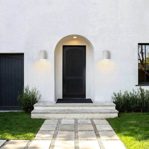 A black arched front door with a small step, flanked by A19 Artisan Lighting’s Leros Downlight Sconces, handcrafted in California, illuminates a white exterior with tiled path and green lawn. LED smart bulbs light the nearby shrubs.