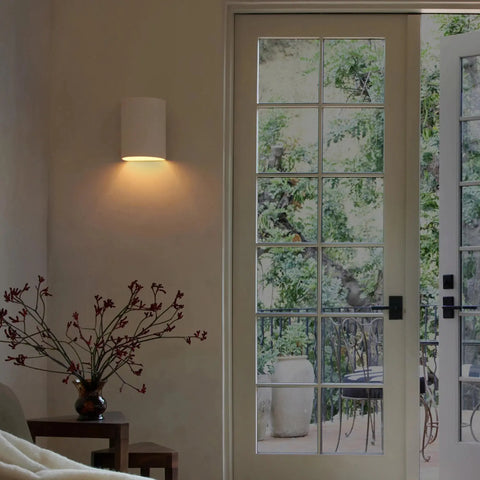 A cozy room features the Leros Downlight Sconce by A19 Artisan Lighting, handcrafted in California, softly illuminating the wall; a small table with red flowers sits nearby, and glass doors open to a patio with greenery and outdoor furniture.