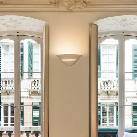 A Modern USA-Made Wall Lighting – Majorca Sconce by A19 Artisan Lighting, handcrafted in California, provides warm LED light between two tall windows with white frames and open shutters, facing a building with green shutters and ornate balconies.