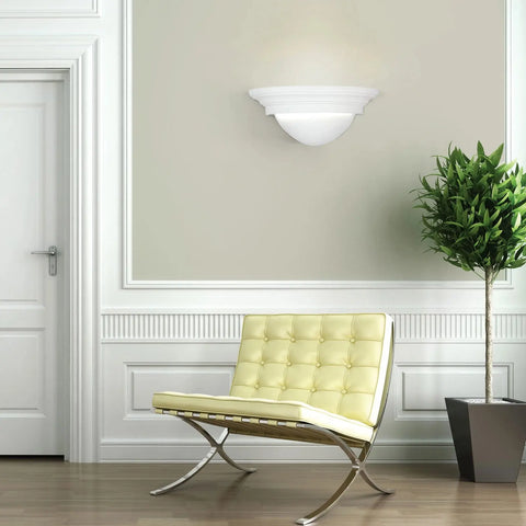 A modern room with a white door, a handcrafted Majorca Sconce by A19 Artisan Lighting, a tall potted plant, and a yellow tufted leather bench with metal frame on wood floors. Light gray walls have white trim and decorative molding.