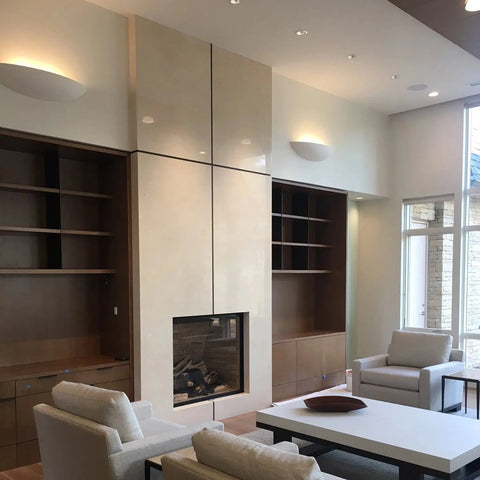 Modern living room with cream walls, built-in wooden shelves, a central fireplace with a beige glossy surround, white armchairs and coffee table, large windows, and inviting Maui Sconce lighting by A19 Artisan Lighting handcrafted in California.
