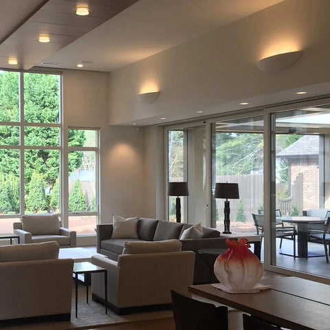 A bright living room features large windows, neutral sofas, a glass coffee table, and patio views. Maui Sconce wall lighting by A19 Artisan Lighting adds handcrafted, modern style. Decorative lamps and a vase with orange accents complete the look.