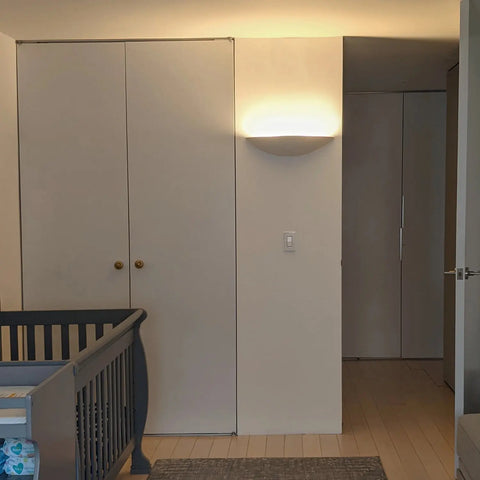 A minimalist nursery featuring a gray crib, double-door closet with gold knobs, light wood floor, textured gray rug, and the Modern USA-Made Wall Lighting – Maui Sconce by A19 Artisan Lighting. Soft LED smart bulbs keep the space uncluttered and serene.