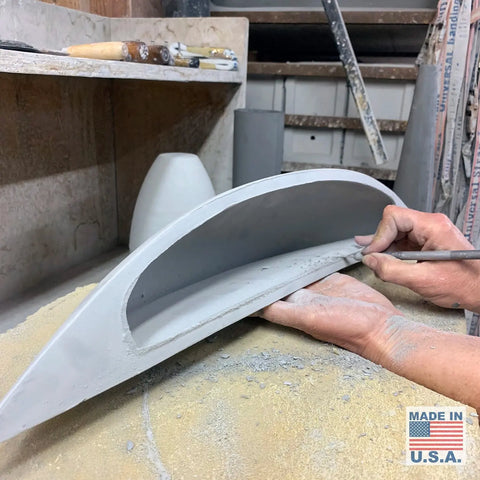 In a workshop filled with tools, an artisan shapes a curved gray sconce. The bottom right displays a Handcrafted in California label for the Modern USA-Made Wall Lighting – Maui Sconce by A19 Artisan Lighting.