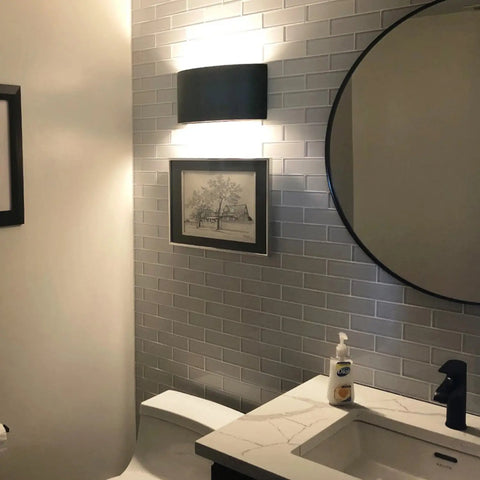 A modern bathroom with gray subway tile walls, a round mirror, and the Modern USA-Made Wall Lighting – Nicosia Sconce by A19 Artisan Lighting above a black-and-white photo. Features include a white toilet and countertop with handmade soap.