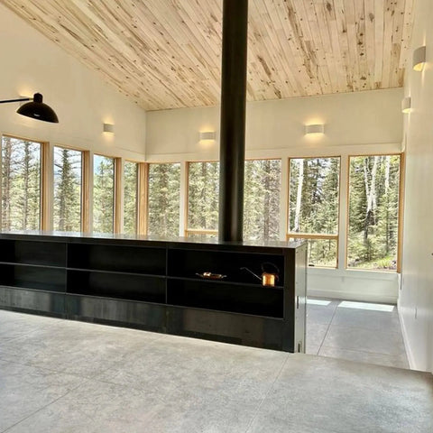 A modern, minimalist room with large windows, light wood ceiling, and black shelving is brightened by the Nicosia Sconce wall lights from A19 Artisan Lighting—handcrafted in California for a fresh, airy feel.