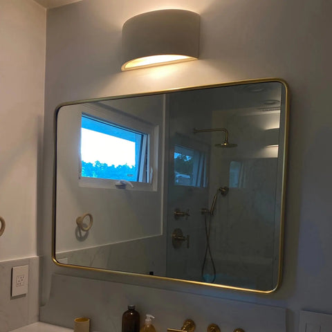 A bathroom mirror with a gold frame reflects the shower and window. Above, an A19 Artisan Lighting Nicosia Sconce—handcrafted in California—casts a warm glow. Toiletries and a towel ring rest on the countertop below.