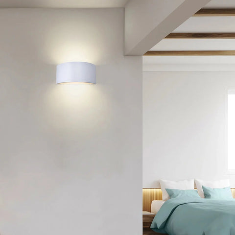 A modern bedroom with teal bedding, a wooden headboard, and a white wall featuring the Nicosia Sconce by A19 Artisan Lighting, handcrafted in California. Natural light from the window enhances the serene ambiance.