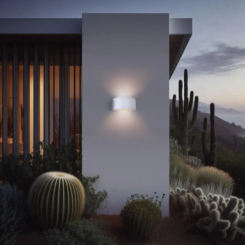 A19 Artisan Lighting’s Modern USA-Made Nicosia Sconce, handcrafted in California, glows on a minimalist homes exterior at dusk amid cacti and desert plants, with mountains in the background.