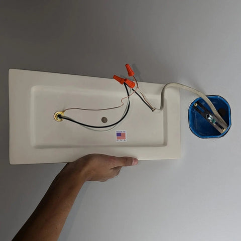 A hand holds the Modern USA-Made Nicosia Sconce by A19 Artisan Lighting, with capped wires near a ceiling and an American flag sticker indicating its handcrafted in California.