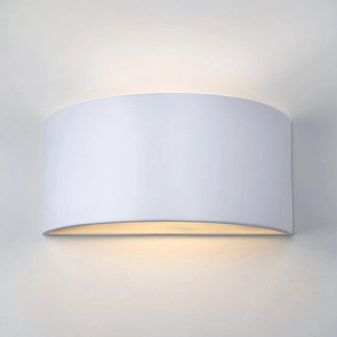 The Modern USA-Made Nicosia Sconce by A19 Artisan Lighting is handcrafted in California, featuring a curved half-cylinder design that emits a soft, warm glow from the top and bottom when mounted on a light-colored wall.