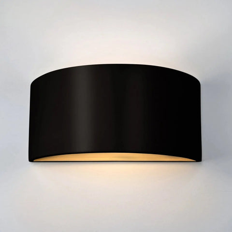 The Modern USA-Made Nicosia Sconce by A19 Artisan Lighting, handcrafted in California, features a curved rectangular black design that shines warm light up and down, shown elegantly on a plain white wall.