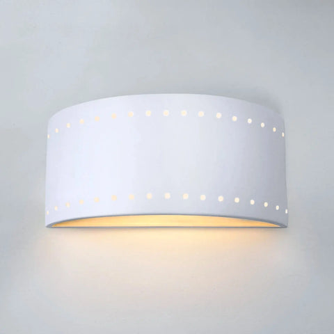 The Paros Downlight Sconce by A19 Artisan Lighting is a modern, handcrafted white wall light with a curved design and circular cutouts, made in California and emitting a soft, warm glow from below.