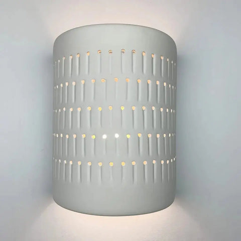 The Samos Sconce by A19 Artisan Lighting is a handcrafted, USA-made cylindrical white wall fixture with vertical rows of small holes that let its LED light create a patterned glow on the wall.