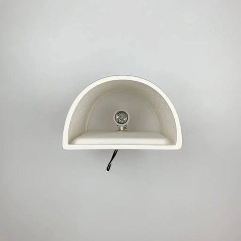 The Samos Sconce by A19 Artisan Lighting is a handcrafted, white ceramic wall light with a semi-circular design, bulb socket, and black cord, shown against a light gray wall. Made in California, USA.