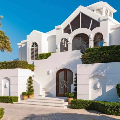 A large, modern white house with arched windows showcases a Modern USA-Made San Marcos Sconce by A19 Artisan Lighting at the grand entrance, surrounded by potted plants, manicured bushes, and stairs leading to wooden double doors against a clear blue sky.