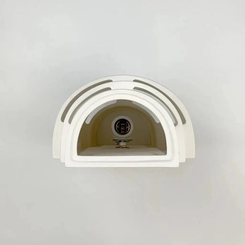 A minimalist white dome with layered arches, inspired by the Modern USA-Made Wall Lighting – San Marcos Sconce from A19 Artisan Lighting, features a central metallic detail and sits on a light surface against a plain gray background. Handcrafted in California.