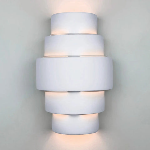 The Modern USA-Made San Marcos Sconce by A19 Artisan Lighting features a handcrafted, tiered cylindrical white design that glows with energy-efficient LED light, adding a warm, contemporary touch to any plain light-colored wall.