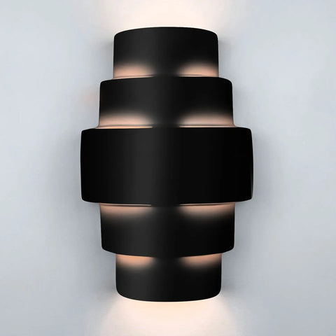 The Modern USA-Made San Marcos Sconce by A19 Artisan Lighting displays a sleek black cylindrical form, casting soft white LED light from multiple points on a light gray wall. Handcrafted in California for energy-efficient illumination.