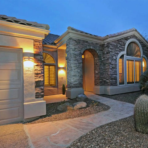 Single-story home with stone and stucco exterior, arched windows with shutters, and warm dusk lighting from A19 Artisan Lighting’s Modern USA-Made Santa Cruz Sconce. A paved walkway leads to the entrance amid desert landscaping with rocks and a cactus.