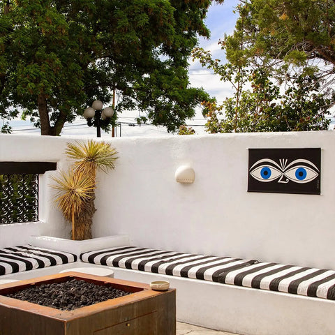 A white patio features black and white benches, a firepit, potted plant, wall art with wide blue eyes, and the Modern USA-Made St Vincent Downlight Sconce by A19 Artisan Lighting above the art. Trees and a partly cloudy sky are in the background.