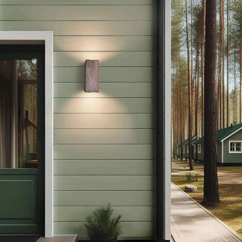 A19 Artisan Lighting’s Modern USA-Made Stone Sconce, handcrafted in California, is mounted on a green wooden house exterior near a glass-paneled door, with pine trees and similar homes visible in the background.