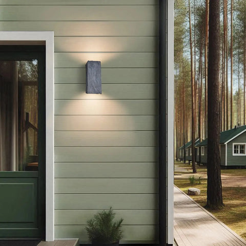 A19 Artisan Lighting’s Modern USA-Made Stone Sconce, handcrafted in California, is mounted by the door on a light green house with white trim; more homes sit among tall pines in the background.