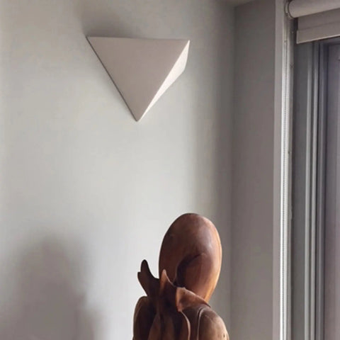 A wooden abstract sculpture stands near a window with blinds, while the A19 Artisan Lighting Sumatra Downlight Sconce—handcrafted in California and featuring energy-efficient LED—illuminates the light gray wall, enhancing the modern aesthetic.
