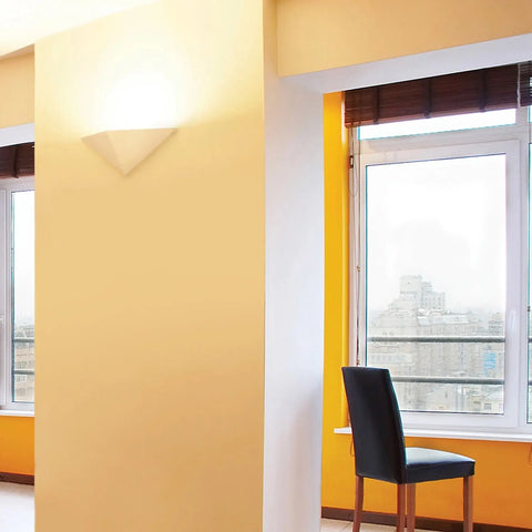 A modern room with yellow walls, a window with brown blinds showing a city view, a black chair by the window, and an A19 Artisan Lighting Sumatra Downlight Sconce—handcrafted in California—glowing on the left wall.